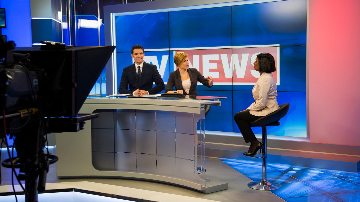A gray news desk at at TV station with a white male anchor, a white female anchor, and a black female guest.