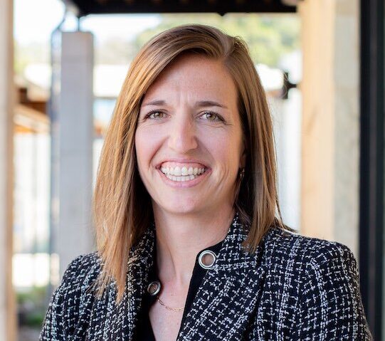 Woman with brown hair, wearing a blue-navy blazer, smiling.