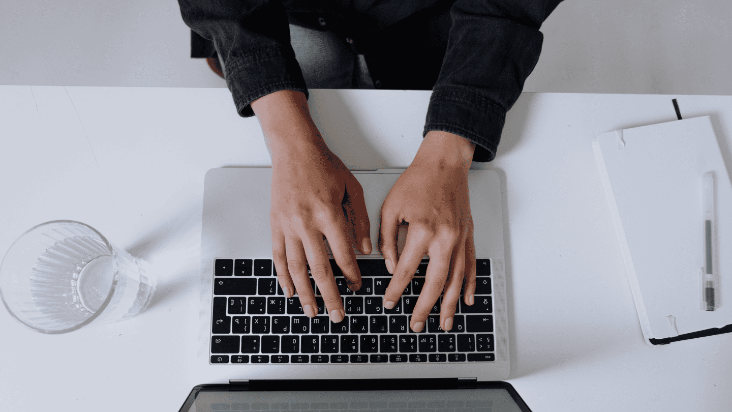 A pair of hands typing on a laptop on a white desk with a clear glass of water on the left and a white journal with a black pen on top of it on the right.