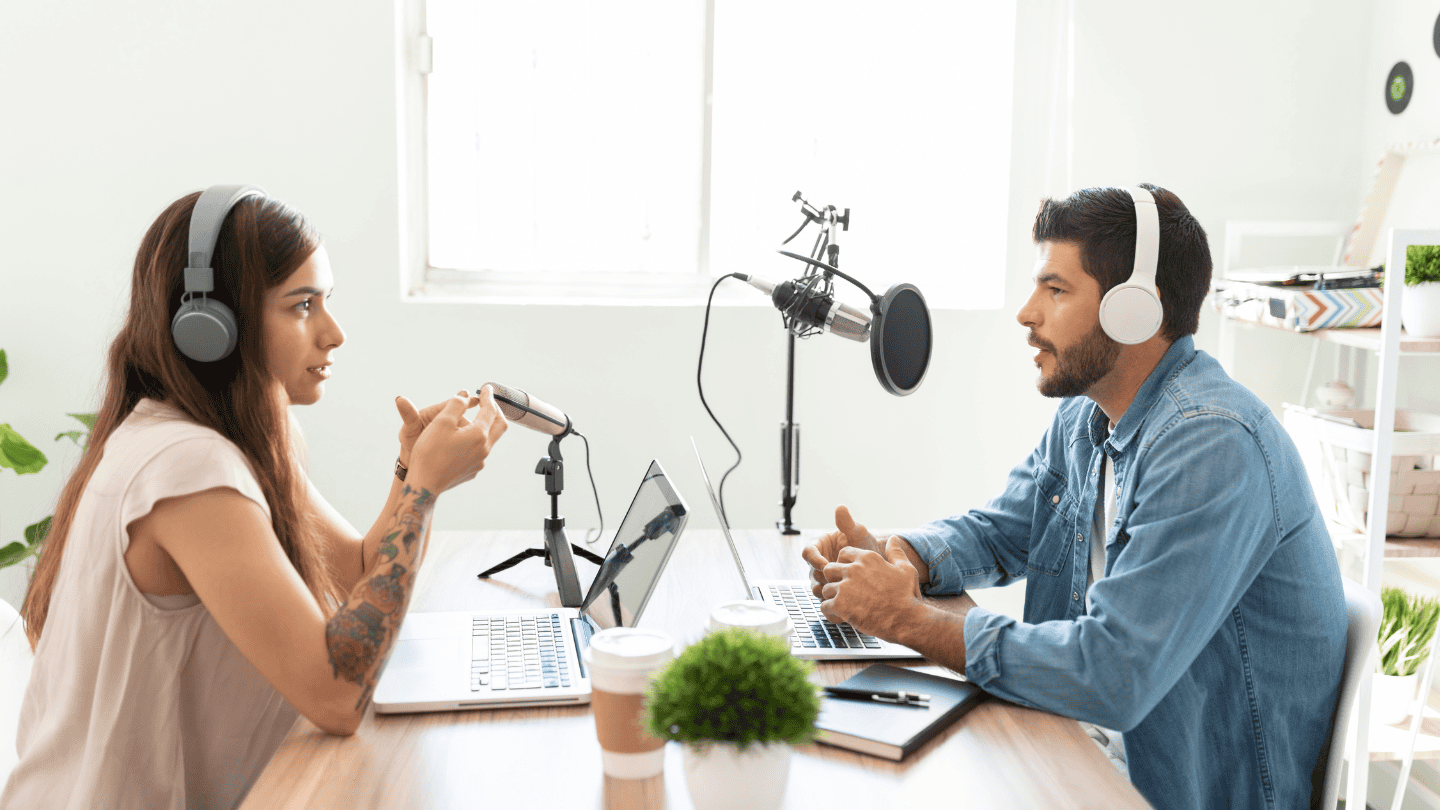 Two people wearing headphones sit across from each other at a table with laptops and microphones, recording a podcast in a bright, casual studio setting.